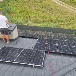 Elektrotechnika Konrad Wykowski - Montaż paneli fotowoltaicznych na ciemnoszarym dachu z dachówki; pracownik w białej koszulce i czapce stoi przy kominie, obok ułożonych modułów, z widocznym zielonym terenem i polną drogą w tle.