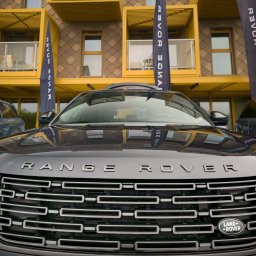 Piotr.Video - Ciemnoszary Range Rover z widocznym grillem i napisem na masce, zaparkowany przed nowoczesnym budynkiem z żółtymi balkonami i flagami marki.