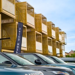 Piotr.Video - Rząd lśniących samochodów Range Rover zaparkowanych przed nowoczesnym budynkiem z ażurowymi, drewnianymi balkonami. Na pierwszym planie maska ciemnego SUV-a z napisem 'RANGE ROVER', w tle błękitne...