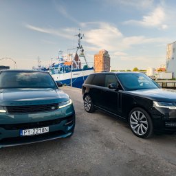 Piotr.Video - Dwa luksusowe SUV-y Range Rover (ciemnozielony i czarny) na asfaltowym nabrzeżu portowym, z dużym statkiem 'RAVEN KOŁ-125' i diabelskim młynem w tle.