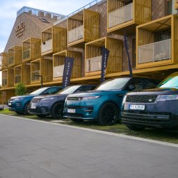 Piotr.Video - Nowoczesny hotel Nickel Resort & Wellness z brązową fasadą i drewnianymi loggiami, przed którym w rzędzie stoją luksusowe SUV-y Range Rover, z flagami promocyjnymi.