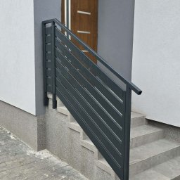 Prywatne - Nowoczesna balustrada z poziomych, antracytowych lameli, zamontowana przy zewnętrznych schodach z jasnych płytek, prowadzących do drewnianych drzwi wejściowych.