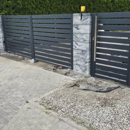 Prywatne - Nowoczesne ogrodzenie z ciemnoszarych, poziomych paneli i bramą, z ozdobnymi słupkami z warstwowego betonu. Widoczny domofon, lampa ostrzegawcza i zielony żywopłot w tle.