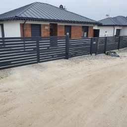 Prywatne - Nowoczesne, ciemnoszare ogrodzenie lamelowe z bramą przesuwną i wbudowaną skrzynką na listy, przed dwoma domami o minimalistycznym designie, na nieutwardzonym podłożu.