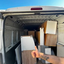 Szybcy I załadowani - Wnętrze białego busa transportowego wypełnione zdemontowanymi szafkami i półkami w odcieniach bieli i drewna, z ręką z tatuażem dającą kciuk w górę, na tle błękitnego nieba.