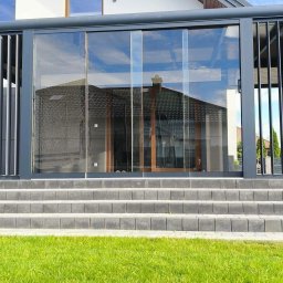 OLAMIS - Zabudowa tarasu z ciemnoszarego aluminium, z przesuwnymi szklanymi drzwiami i pionowymi lamelami, prowadzą do niej kamienne schody.