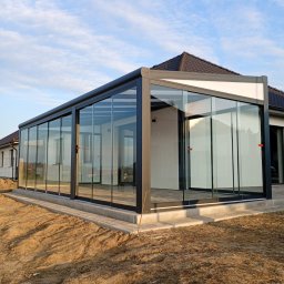 OLAMIS - Nowoczesna szklana zabudowa tarasu z ciemną, metalową ramą, przylegająca do białego domu z brązowym dachem, na betonowej podmurówce, otoczona piaszczystym terenem pod błękitnym niebem.