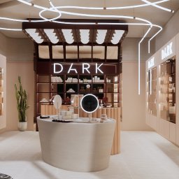 MERIDIAN EU Sp. z o. o. - Nowoczesne wnętrze showroomu z owalnym kontuarem ekspozycyjnym, podświetlanym logo 'DARK' i unikalnym, liniowym oświetleniem sufitowym. Dominują ciepłe beże i brązy, z lustrzanymi akcentami.