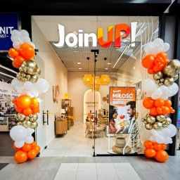 MERIDIAN EU Sp. z o. o. - Ukończony salon Join UP! z przeszklonym wejściem, udekorowany pomarańczowo-biało-złotymi girlandami balonowymi. Wnętrze z drewnianymi meblami, plecionymi lampami i jasnymi płytkami.