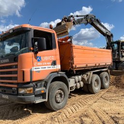 Dariusz Kubicz - Pomarańczowa ciężarówka Scania 124c 470 firmy KUBICZ jest ładowana jasnym urobkiem przez koparkę Volvo na placu budowy. W tle błękitne niebo z chmurami.