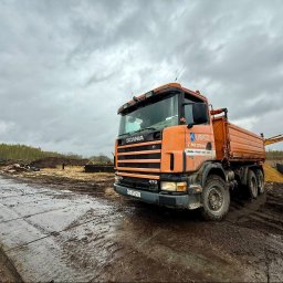 Dariusz Kubicz - Pomarańczowa wywrotka Scania firmy UBICZ na błotnistym placu budowy, z widocznymi panelami drogowymi i hałdami ziemi w tle, pod pochmurnym niebem.