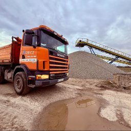 Dariusz Kubicz - Pomarańczowa wywrotka Scania firmy KUBICZ z widocznym numerem telefonu, zaparkowana na błotnistym placu. W tle duża hałda kruszywa i żółty przenośnik taśmowy pod zachmurzonym niebem.