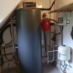 Schelinger - Polski producent pomp ciepła i zbiorników buforowych 12 lat gwarancji - Ciemnoszary zbiornik buforowy z rozbudowaną instalacją rur, zaworów i dwoma naczyniami wzbiorczymi w kotłowni. Na zbiorniku leży pudełko filtra magnetycznego IBO.