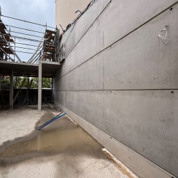 FIRMA Budownictwo - Surowa betonowa ściana z wyraźnymi poziomymi łączeniami, obok rusztowania i betonowego stropu na placu budowy, z kałużą na podłożu i wystającymi przewodami.