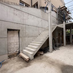 FIRMA Budownictwo - Betonowe schody zewnętrzne i masywna ściana oporowa w trakcie budowy, z widocznymi śladami szalunku i zbrojeniem. W tle beżowy budynek i rusztowania.