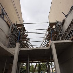 FIRMA Budownictwo - Budowa żelbetowej konstrukcji z betonowymi słupami i płytą, widoczne metalowe rusztowania i szalunek, w przestrzeni między dwoma beżowymi budynkami.