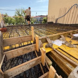 FIRMA Budownictwo - Przygotowanie zbrojenia stropu betonowego na żółtych szalunkach, z widocznym pracownikiem, planami budowy i wystającą niebieską rurą. W tle miejski krajobraz.