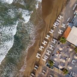 Bartłomiej Bania Net Solutions - Z lotu ptaka: piaszczysta plaża z rozbijającymi się falami, przylegający do niej kurort z basenem, rzędami leżaków, parasolami i palmami, widoczna mała łódź na tarasie.