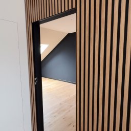 Paweł Jurkowski Wykończenia Wnętrz Premium - Nowoczesne wnętrze z drewnianymi panelami ściennymi w formie pionowych listew, czarną ościeżnicą i jasną podłogą. W tle pokój z ciemnymi ścianami i oknem dachowym.
