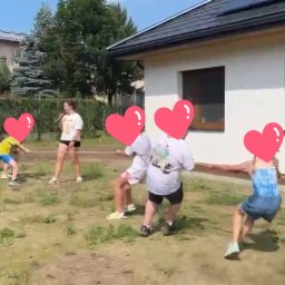 Chichralnia - Grupa dzieci i młodzieży z zamazanymi twarzami intensywnie przeciąga linę na słonecznym, trawiastym podwórku obok nowoczesnego domu z panelami słonecznymi.