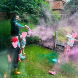 Chichralnia - Kobieta z niebieską farbą na ramieniu rozpyla różowy proszek Holi, a troje dzieci bawi się na zielonym trawniku z kolorowymi plamami. W tle gęsta roślinność i dom.
