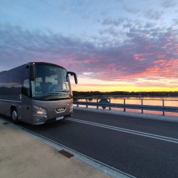 Baltic TRAVEL - Nowoczesny, szary autokar VDL na moście, oświetlony promieniami zachodzącego słońca, z jeziorem i malowniczym niebem w tle.