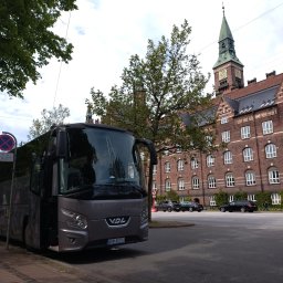 Baltic TRAVEL - Nowoczesny, szary autokar VDL z otwartymi drzwiami, zaparkowany przy ulicy w mieście, z zabytkowym ceglanym ratuszem z zieloną wieżą zegarową w tle. Polska rejestracja.