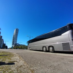 Baltic TRAVEL - Szary autokar turystyczny zaparkowany na tle błękitnego nieba i nowoczesnych budynków, w tym charakterystycznego wieżowca Turning Torso w Malmö. Ujęcie z niskiej perspektywy.