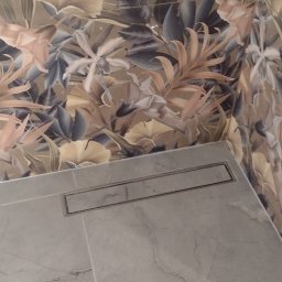 Usługi ogulnobudowlane - Wnętrze z beżową tapetą w botaniczny wzór liści i kwiatów, połączoną z jasnoszarymi płytkami imitującymi marmur, z widocznym liniowym odpływem podłogowym.