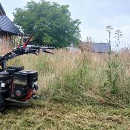 ArborAlp - Ciężka kosiarka bijakowa na niezagospodarowanej działce z wysoką trawą, obok domu w budowie z cegły i czarnym dachem. Część terenu już oczyszczona.