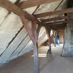 Poddasze z drewnianą konstrukcją więźby dachowej, ocieplone pianką natryskową, widoczna podłoga z płyt OSB i fragment muru z cegły, w tle sylwetka osoby.