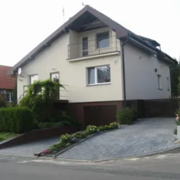 Elewacja domu jednorodzinnego z balkonem, po renowacji. Jasny tynk, brązowe elementy wykończeniowe, zadbany ogród z kwiatami i kostką brukową na podjeździe.