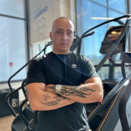 Mikołaj Wachowiak - Łysy mężczyzna z niebieskimi oczami i tatuażami na przedramionach, w czarnej koszulce Adidas, stoi z założonymi rękami w jasnej siłowni z maszynami cardio i dużymi oknami.