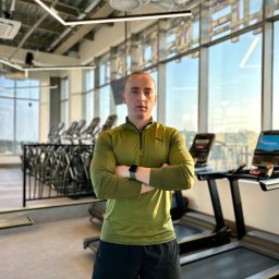 Mikołaj Wachowiak - Mężczyzna w oliwkowej bluzie sportowej i czarnych szortach, z rękami skrzyżowanymi, stoi w nowoczesnej siłowni z dużymi oknami i bieżniami w tle.