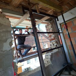 BAU-PERSONAL TADEUSZ SZTONCEL - Uśmiechnięty pracownik w czarnej kurtce i czerwonych rękawicach pozuje na metalowym rusztowaniu, w otworze ściany remontowanego budynku z widocznymi cegłami i belkami stropowymi.