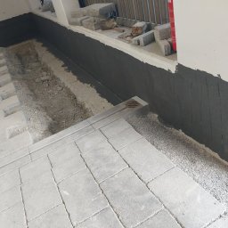 BAU-PERSONAL TADEUSZ SZTONCEL - Ciemna masa bitumiczna na ścianie fundamentowej, obok której widoczny jest głęboki wykop drenażowy oraz częściowo ułożona jasnoszara kostka brukowa z okrągłym odpływem.