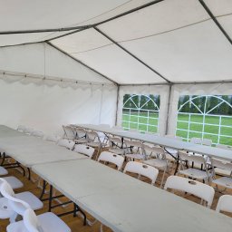Paweł Jamróz Wynajem Atrakcji Okolicznościowych - Wnętrze białego namiotu eventowego z rzędami szarych składanych stołów i białych krzeseł, z widokiem na zielony trawnik przez łukowe okna.