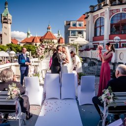 Sieczyńscy Fotograf i Kamerzysta Ślubny - Patryk Sieczyński - Plenerowy ślub w Sopocie: para młoda całuje się pod kwiatową bramą, w tle latarnia morska i zabytkowy budynek. Goście siedzą na krzesłach, świadkowa rzuca bukiet.