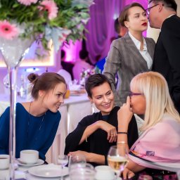 Sieczyńscy Fotograf i Kamerzysta Ślubny - Patryk Sieczyński - Uśmiechnięte kobiety przy elegancko zastawionym stole z wysokim wazonem pełnym różowych gerber. W tle para w garniturze i szarym kostiumie.