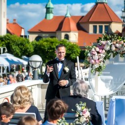 Sieczyńscy Fotograf i Kamerzysta Ślubny - Patryk Sieczyński - Elegancki mężczyzna w smokingu przemawiający podczas uroczystości plenerowej w Otominie, z gośćmi i bogatą dekoracją kwiatową, na tle historycznej architektury.