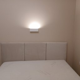 Elektro-Smart - Nowoczesna lampa ścienna zamontowana nad beżowym zagłówkiem łóżka, oświetlająca ścianę rozproszonym światłem. Widoczne dwa białe włączniki światła po bokach łóżka.