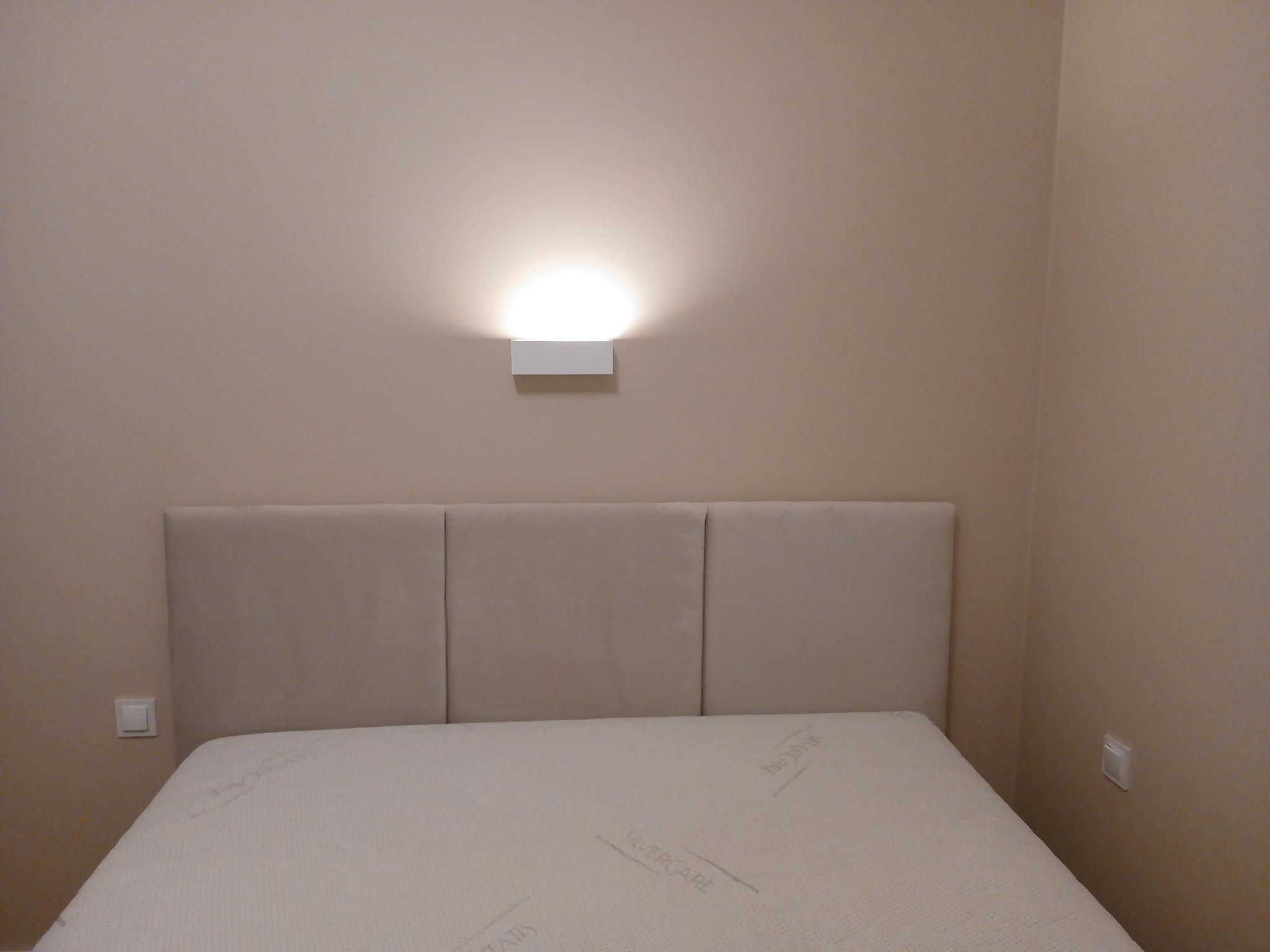 Nowoczesna lampa ścienna zamontowana nad beżowym zagłówkiem łóżka, oświetlająca ścianę rozproszonym światłem. Widoczne dwa białe włączniki światła po bokach łóżka.