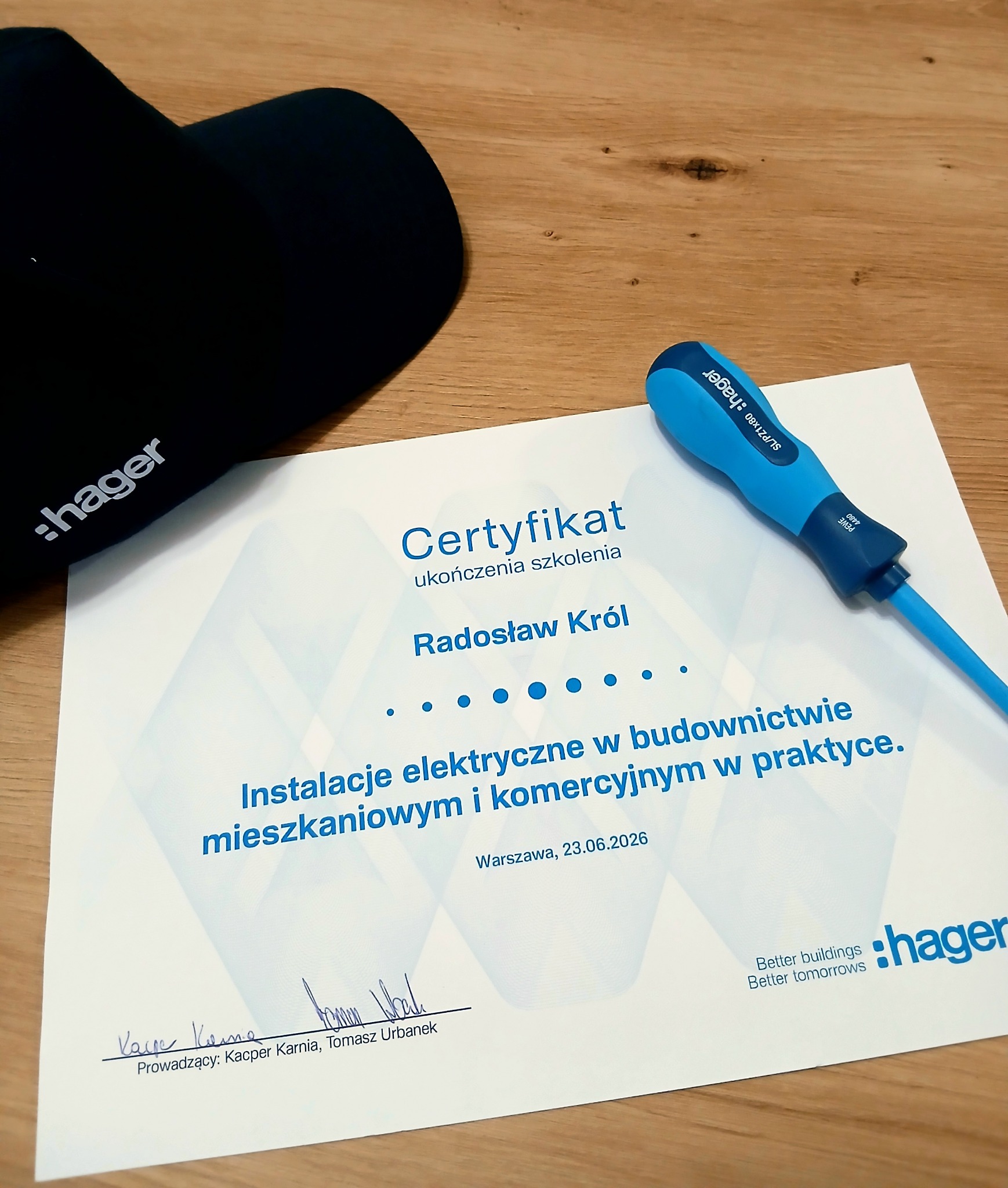 Certyfikat ukończenia szkolenia 'Instalacje elektryczne w budownictwie' dla Radosława Króla, z datą 23.06.2026, na drewnianym tle z czapką i niebieskim śrubokrętem marki Hager.