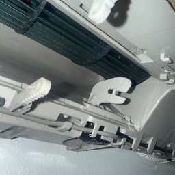 KlimFaktor - Wnętrze jednostki klimatyzacyjnej z widocznym ciemnym, zabrudzonym wentylatorem i białymi elementami mechanizmu sterującego nawiewem. Ujęcie z bliska, od dołu, ukazujące stan przed czyszczeniem.