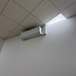 KlimFaktor - Biały klimatyzator ścienny zamontowany pod sufitem podwieszanym z kasetonów, z widocznym korytkiem instalacyjnym i jasnym światłem z wnęki.
