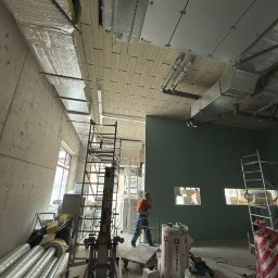 KlimFaktor - Montaż izolowanych kanałów wentylacyjnych i paneli sufitowych w surowym wnętrzu biurowca, z widocznymi rusztowaniami i pracownikiem na etapie instalacji.