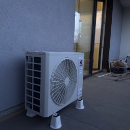KlimFaktor - Zainstalowana jednostka zewnętrzna klimatyzacji GREE na tarasie z szarych płytek, wsparta na antywibracyjnych podstawkach, przy teksturowanej ścianie budynku.