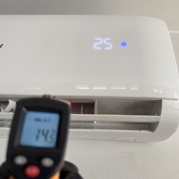 KlimFaktor - Biała klimatyzacja z cyfrowym wyświetlaczem '25', gdzie ręka w czarnej rękawiczce mierzy temperaturę wylotową 14.3°C termometrem, weryfikując jej wydajność.