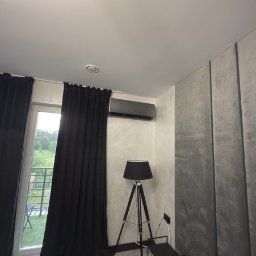 KlimFaktor - Nowoczesna sypialnia z zamontowaną na ścianie czarną jednostką klimatyzacji, ciemnymi zasłonami przy oknie i lampą podłogową na trójnogu, obok ściany z efektem betonu.
