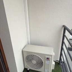 KlimFaktor - Zewnętrzna jednostka klimatyzacyjna GREE, zamontowana w rogu balkonu wyłożonego sztuczną trawą, obok białej ściany i ciemnej balustrady.